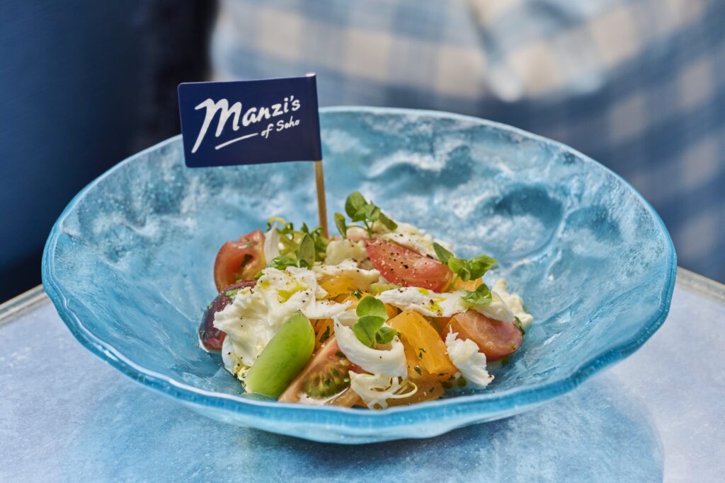 A vibrant Isle of Wight heritage tomato salad with buffalo mozzarella, fresh basil, and microgreens, drizzled with olive oil and black pepper, served in a textured blue glass bowl with a Manzi’s of Soho flag.