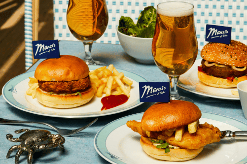 Three plated burgers from Manzi’s Soho, including a fish burger with chips inside the bun, served with fries and glasses of beer on a blue table, with small Manzi’s flags in each burger