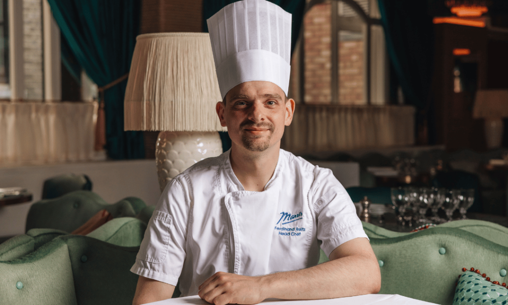 A head shot of a male chef in a white chef jacket and toque hat in a green resplendent restaurant with a lamp behind him